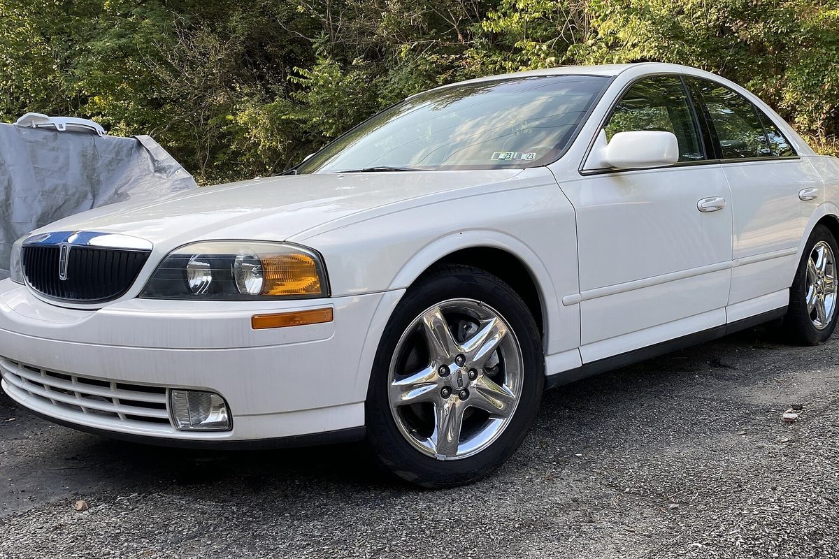 A white 2002 Lincoln LS sedan on a gravel lot next to a car under a gray cover, with trees behind