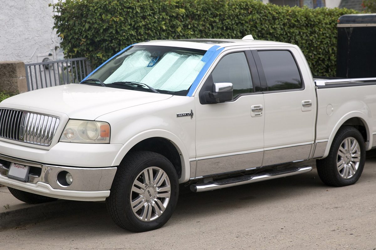 A white Lincoln Mark LT crew-cab pickup with a sunshade in the windshield, parked on a street in front of a tall hedge