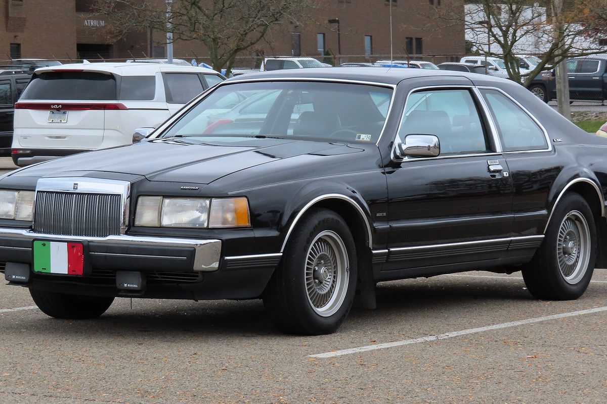 A black Lincoln Mark VII two-door coupe with wire-style wheel covers, parked in a lot beside a white minivan