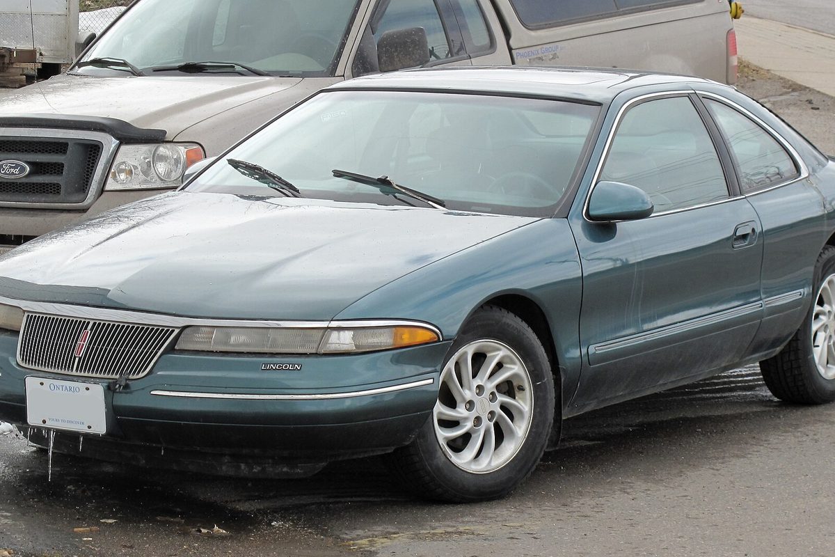 A dark green Lincoln Mark VIII coupe parked on wet pavement next to a silver pickup truck
