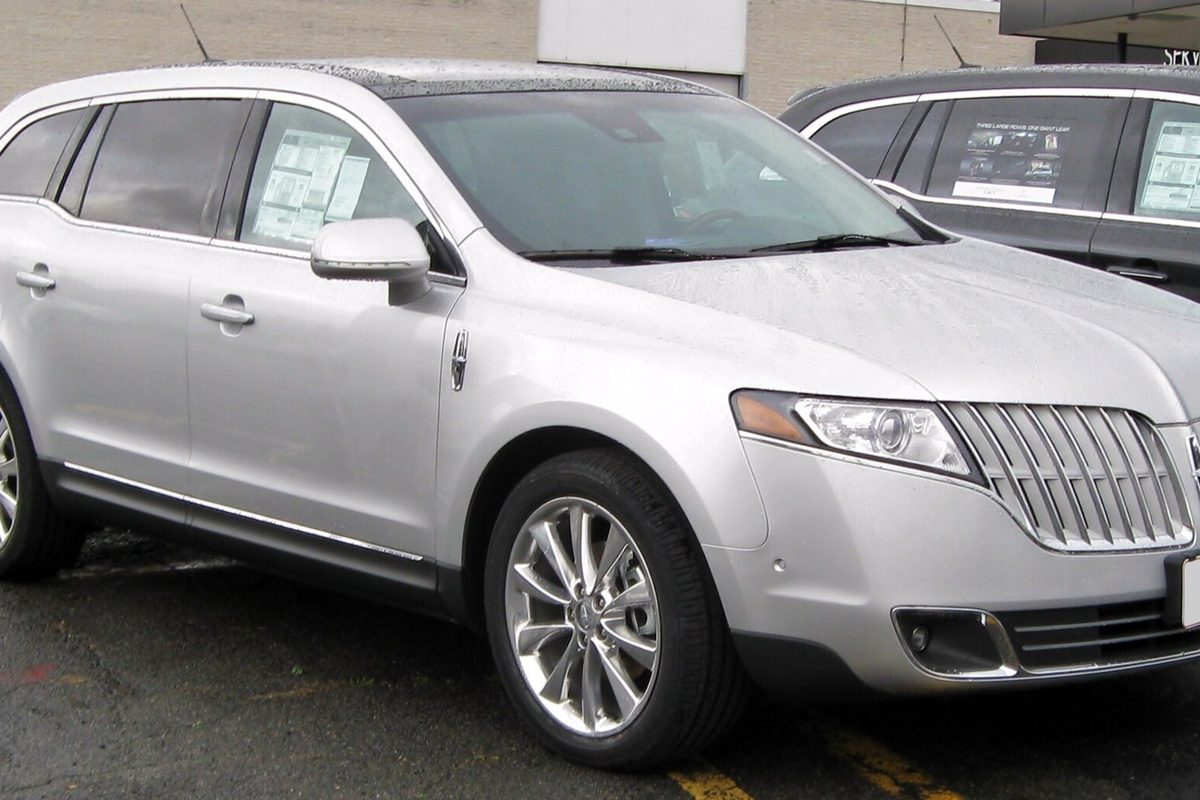 A silver Lincoln MKT crossover with a wide vertical-bar grille, parked on wet pavement with stickers in its windows