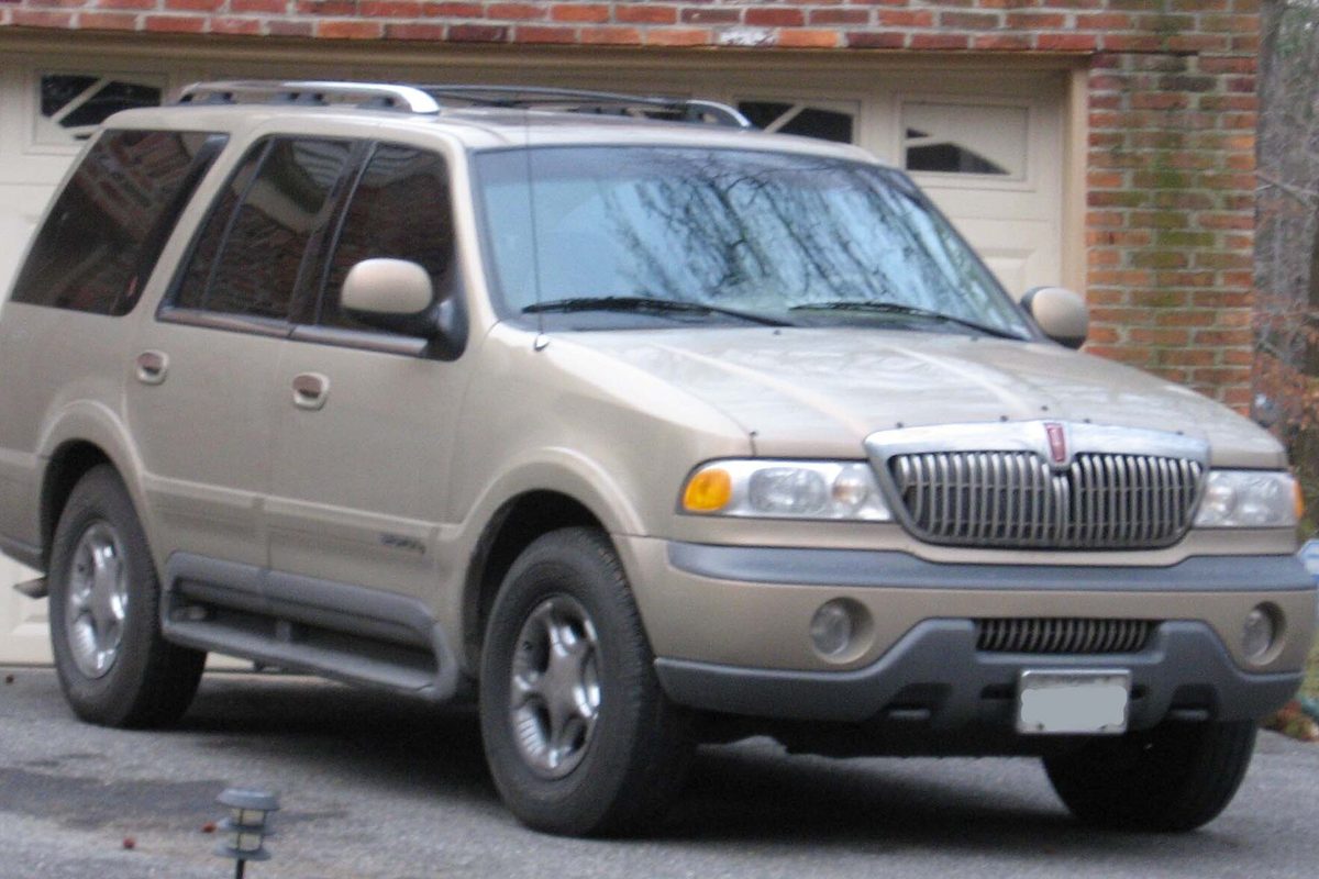 A tan first-generation Lincoln Navigator SUV with a roof rack, parked in a driveway in front of a brick garage