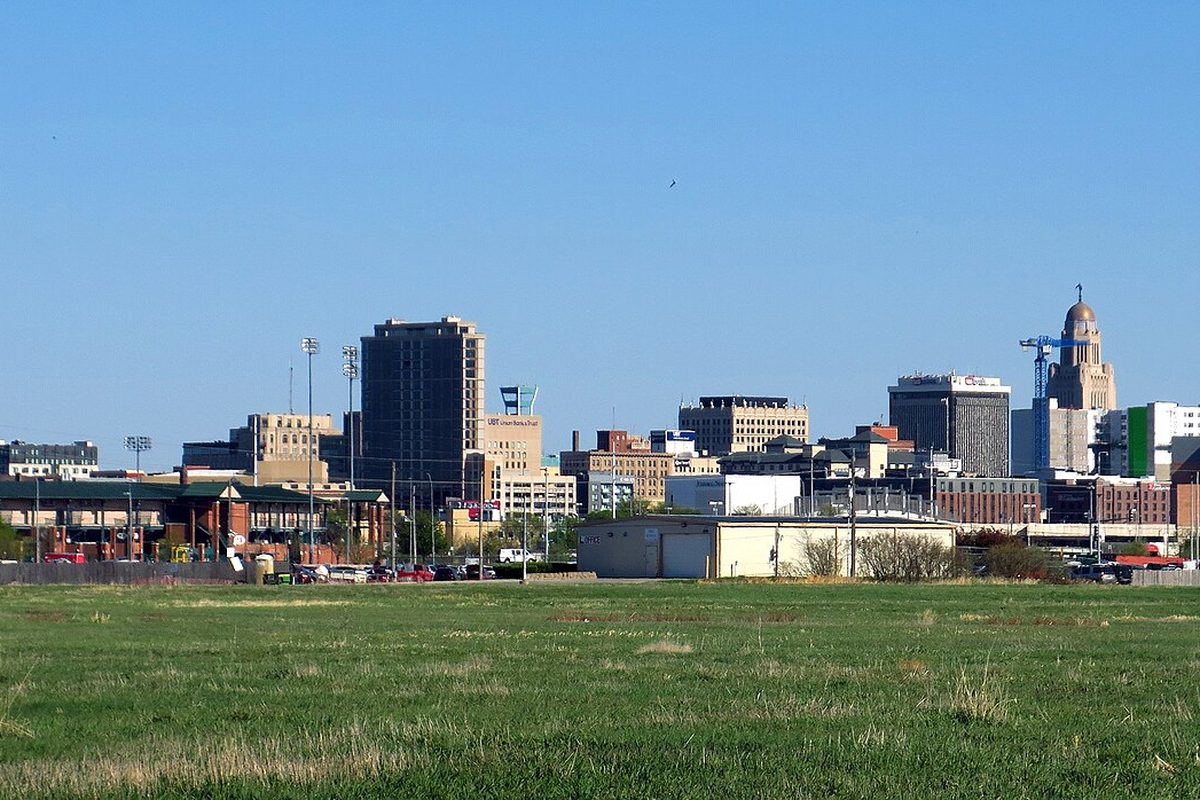 The downtown Lincoln skyline seen across a wide green field, with office towers in the middle, a tall domed tower at right and ballpark light poles at left under a clear sky