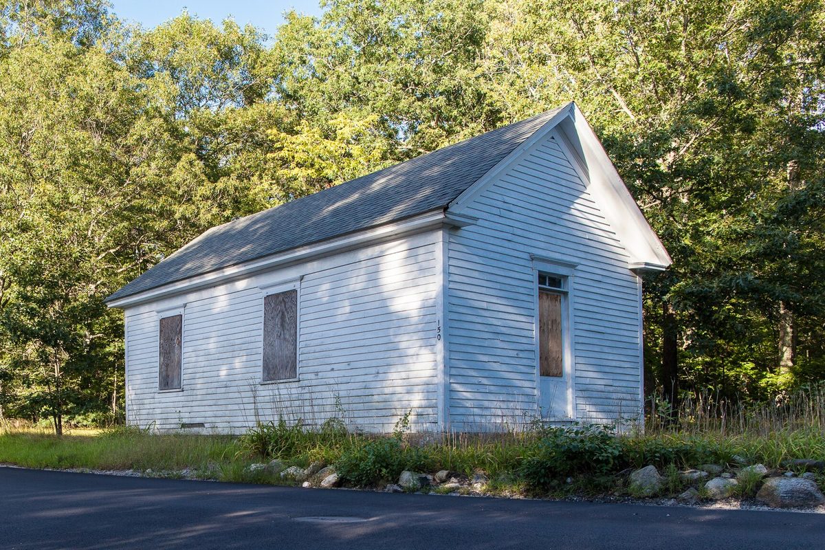 The small white clapboard Pullen Corner School, a one-room schoolhouse with boarded-up windows, among green trees beside a paved road in Lincoln