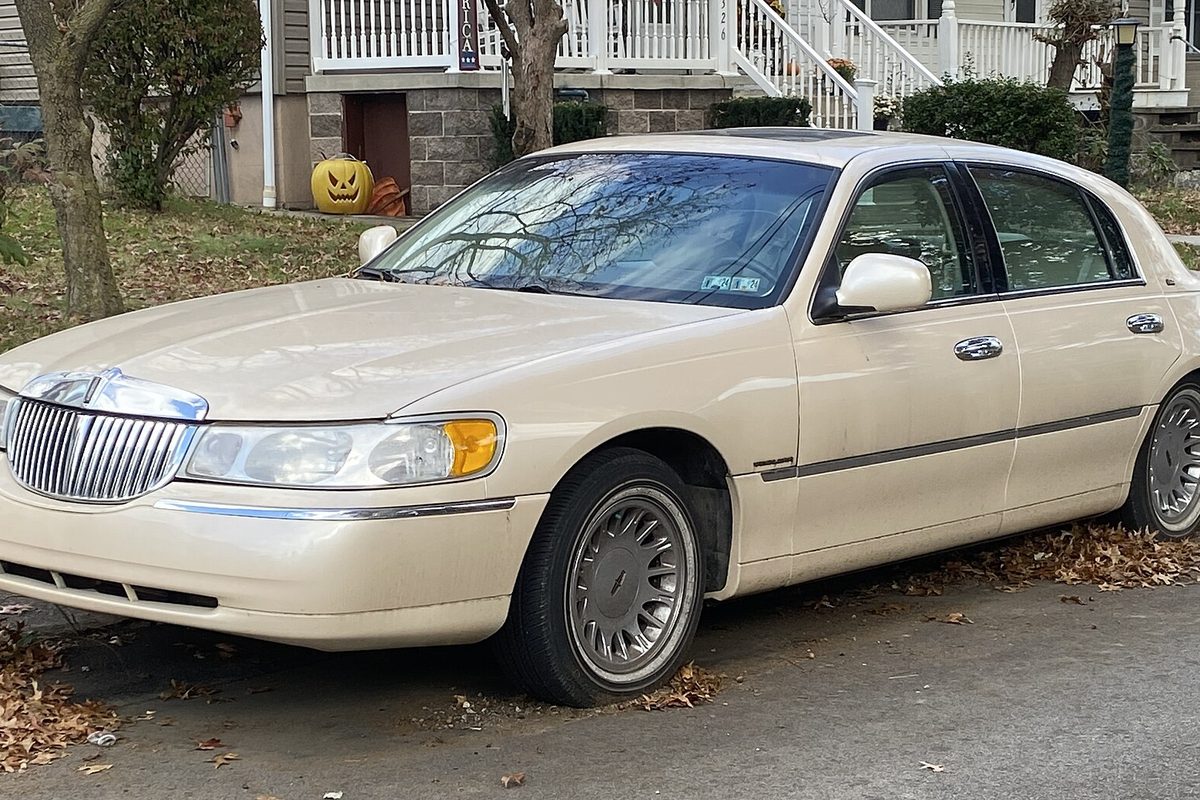 An ivory 1998 Lincoln Town Car sedan parked at a curb in fallen leaves, with a carved pumpkin on the porch steps behind it