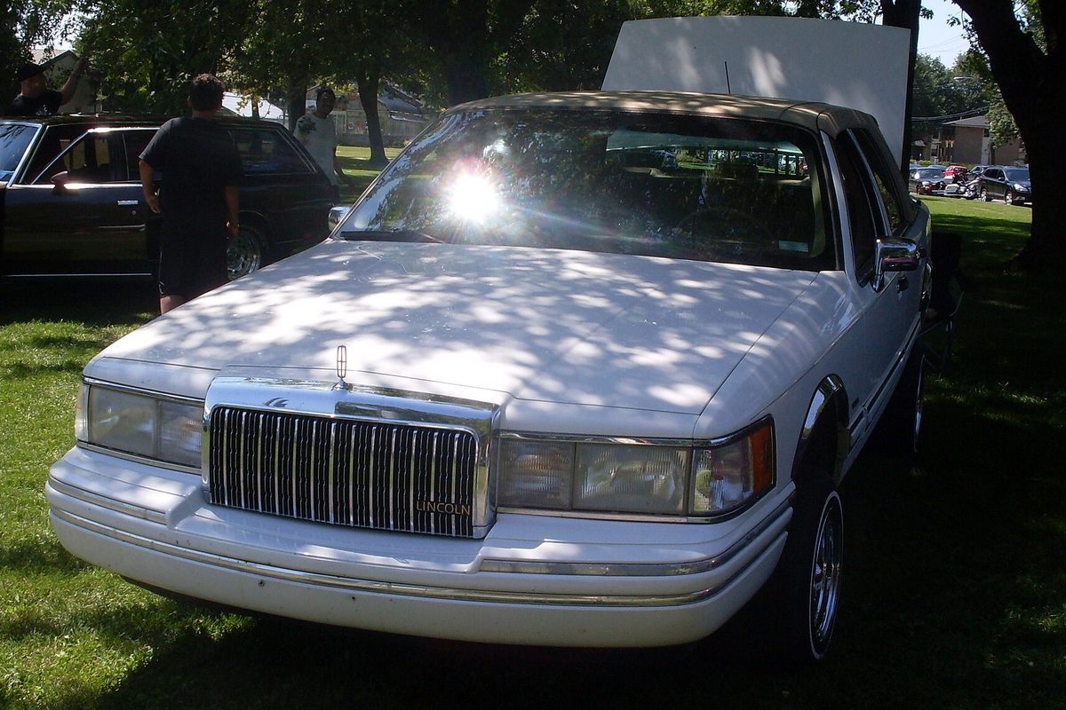 A white 1993-1994 Lincoln Town Car sedan parked on the grass under shade trees at a lowrider club picnic in Montreal