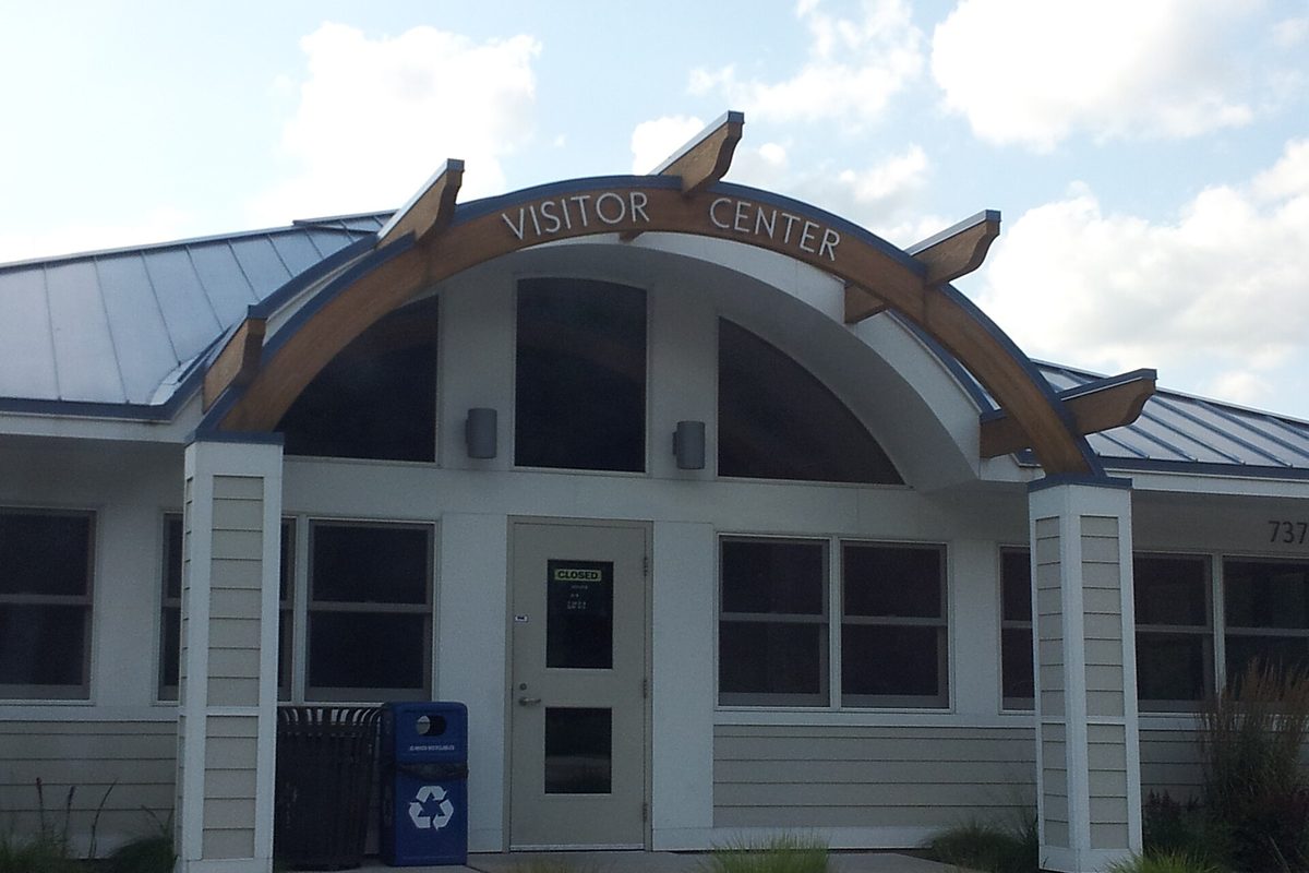 The Lino Lakes Visitor Center, a light gray building with a metal roof and a curved wooden arch over the entrance reading Visitor Center, with a blue recycling bin beside the door