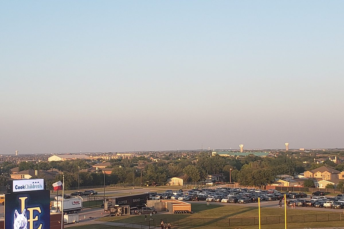View from the Little Elm athletic complex across a full parking lot and tree-lined rooftops toward two water towers, with the school's LE sign in the corner