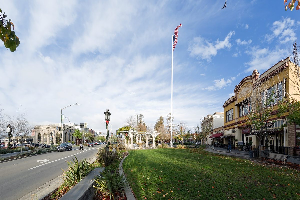 Downtown Livermore's tall flagpole on a green lawn, with an old-style street clock, a white pergola and storefronts under a partly cloudy sky