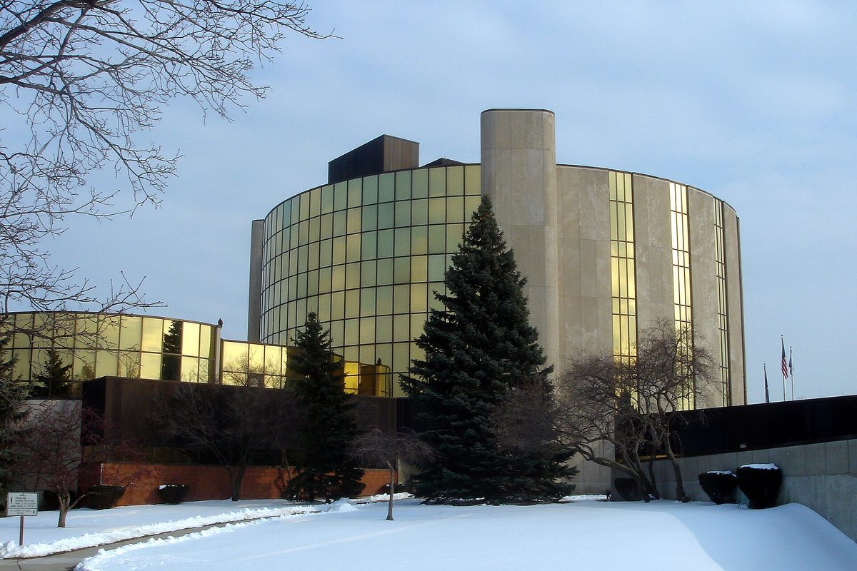 Livonia City Hall, a round tower of gold-tinted glass and pale concrete behind tall evergreens, with snow covering the lawn