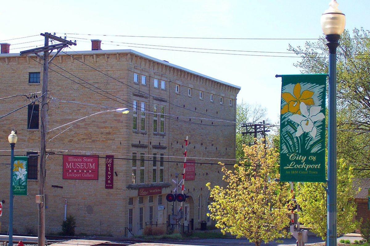 A three-story limestone building in Lockport's historic district with an Illinois State Museum banner, a railroad crossing signal and a green City of Lockport banner on a lamppost