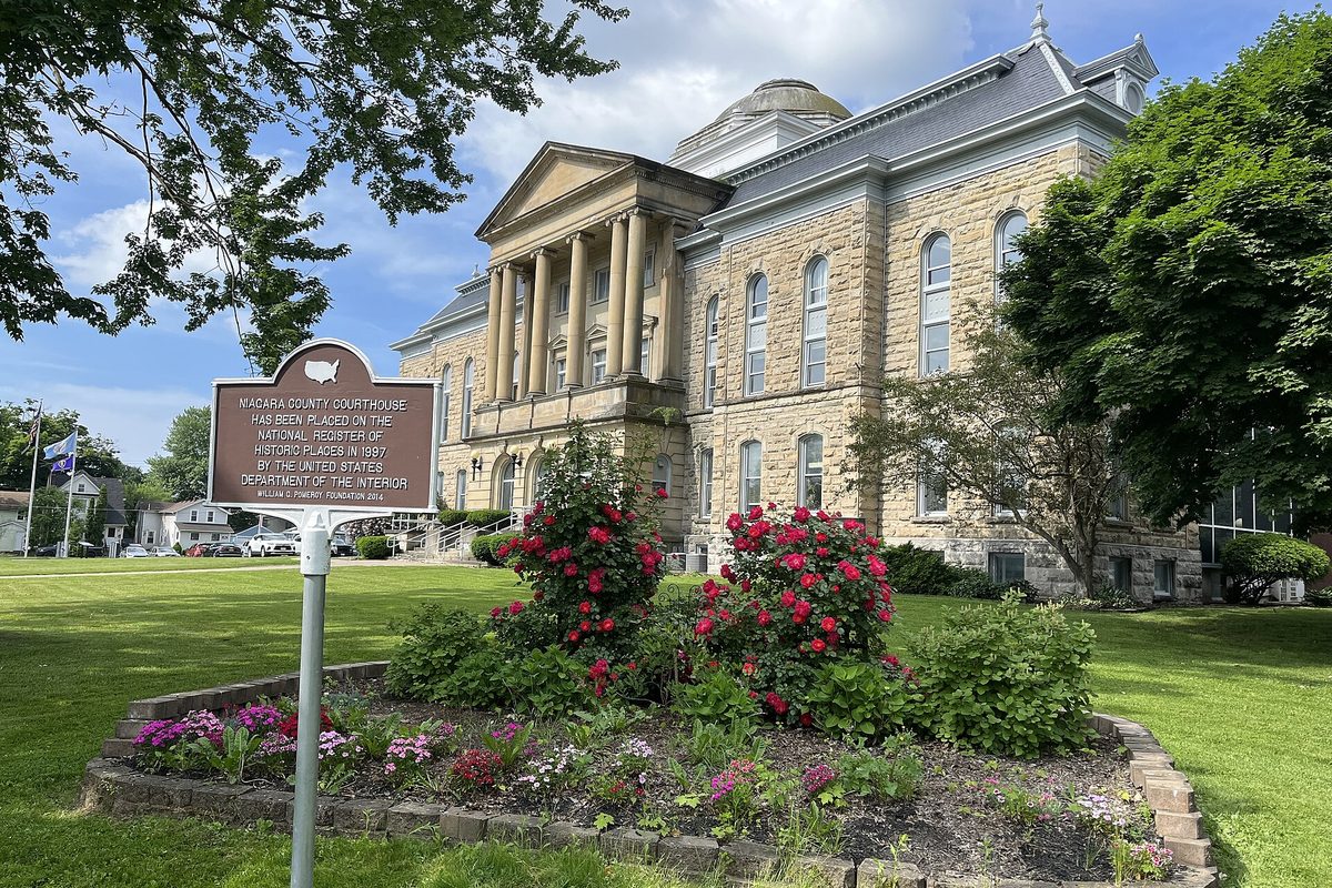 The domed stone Niagara County Courthouse behind a bed of red roses, with a historic marker sign on the lawn in front
