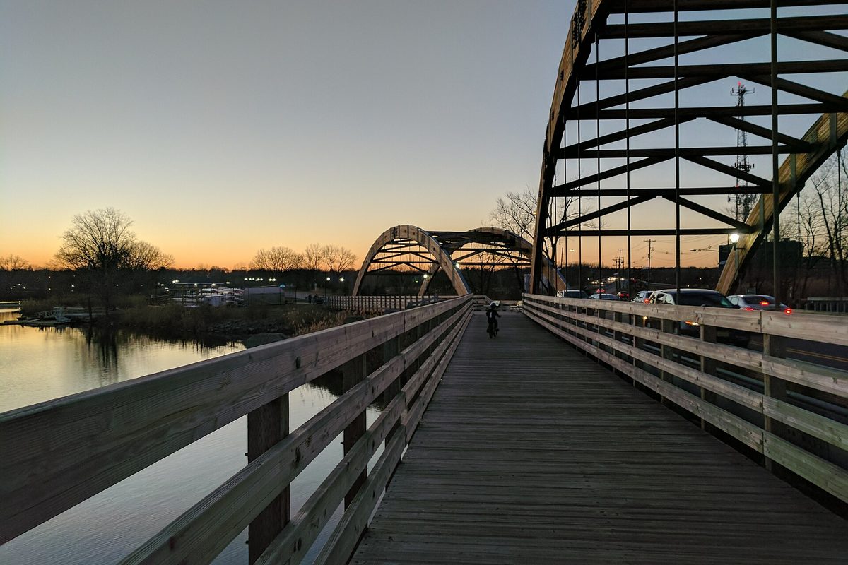 A wooden footbridge under arched steel trusses at sunset in Overpeck County Park, with still water on the left and a cyclist on the deck
