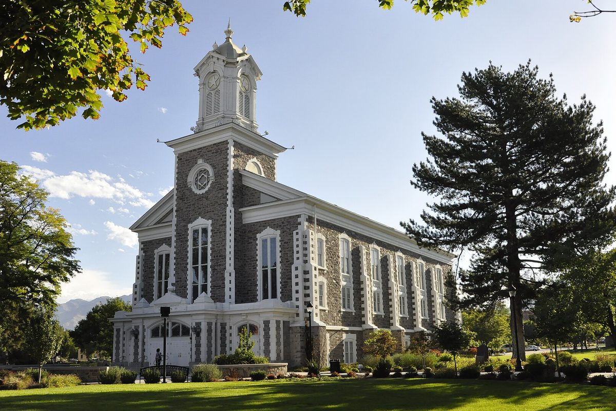 The gray stone Logan Tabernacle with a white clock tower and trim, on a green lawn beside a tall pine under a blue sky