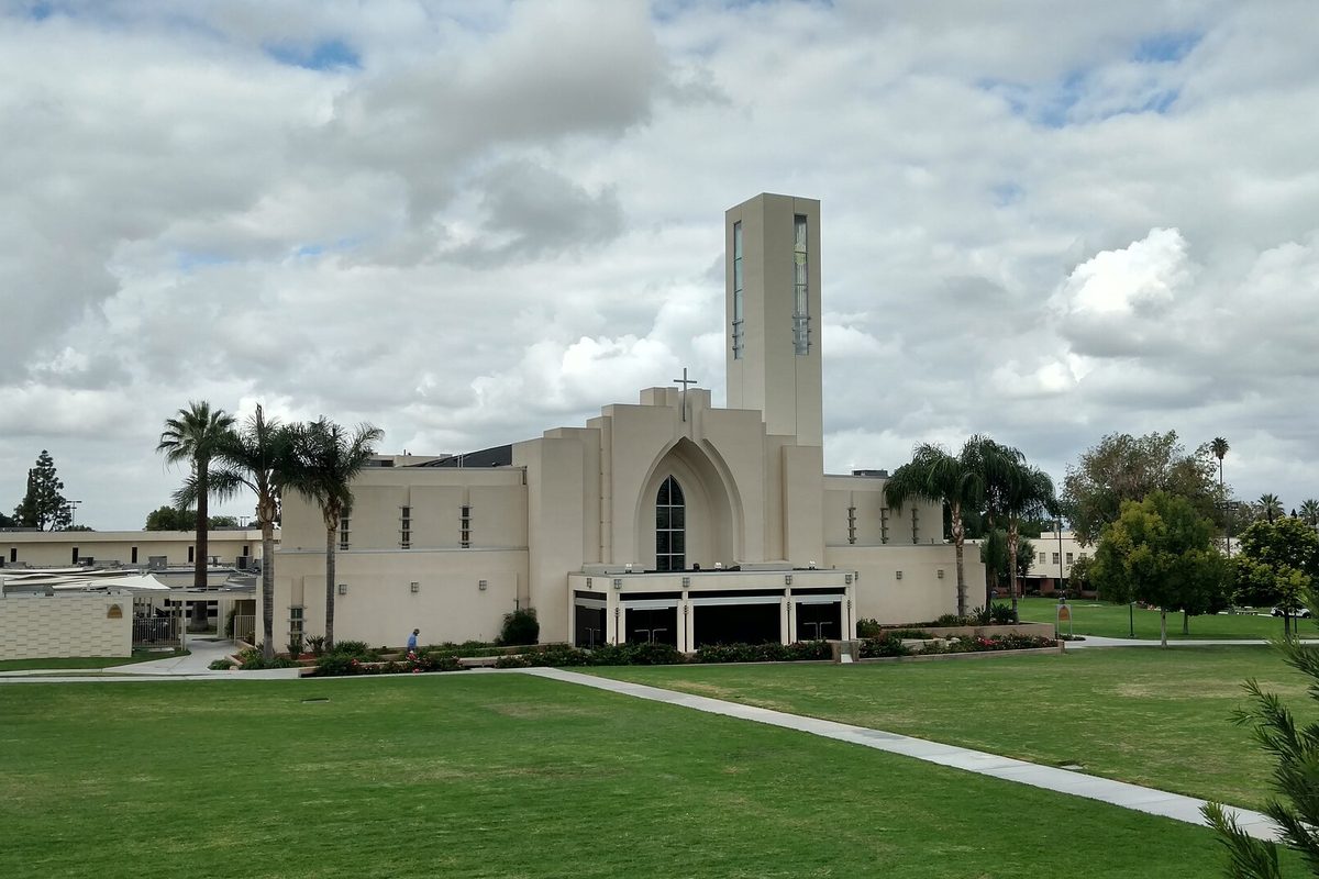 A white modern chapel with a tall square tower and a cross, palm trees and a wide green lawn under a cloudy sky in Loma Linda