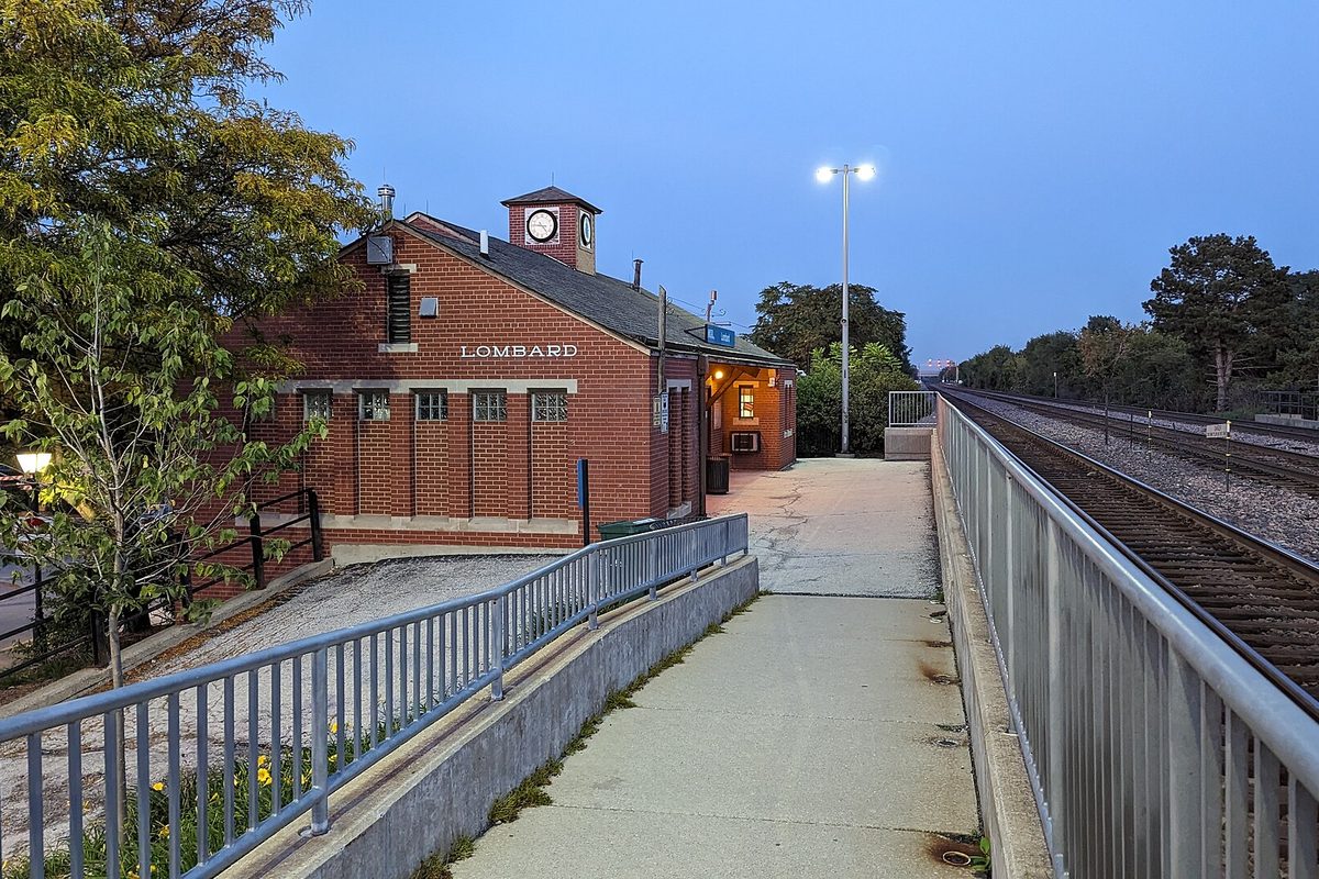 The red brick Lombard Metra station building with a clock cupola and lit windows beside a railed walkway and the tracks at dusk