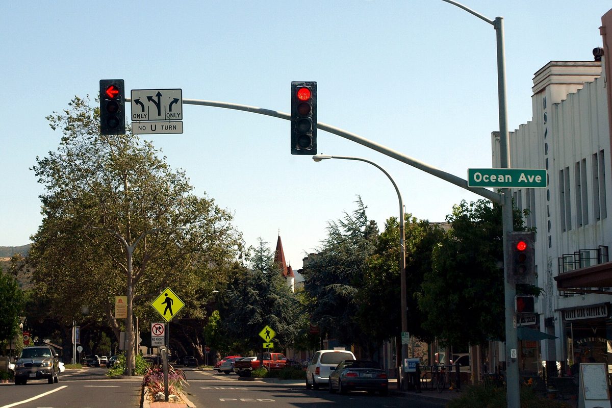Traffic lights and an Ocean Ave street sign over downtown Lompoc, with trees, parked cars and a white art deco building on the right