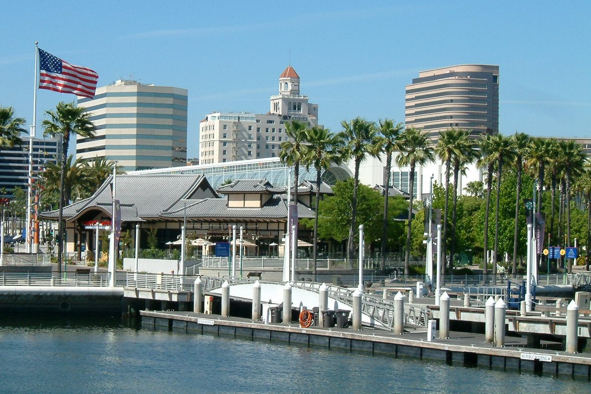 The Long Beach waterfront with a pagoda-roofed building, palm trees, an American flag and downtown towers behind a marina dock