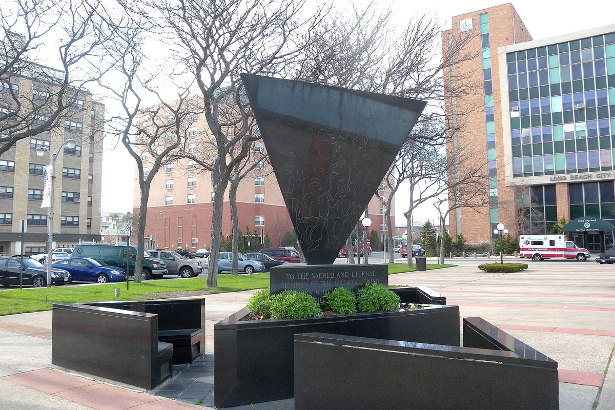 A dark inverted-triangle Holocaust memorial on a plaza in front of Long Beach City Hall, with bare trees, apartment towers and an ambulance parked nearby
