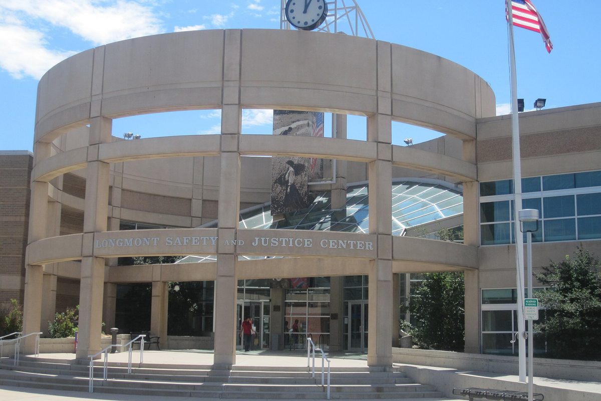 The curved stone colonnade of the Longmont Safety and Justice Center, with a clock tower and an American flag against a blue sky