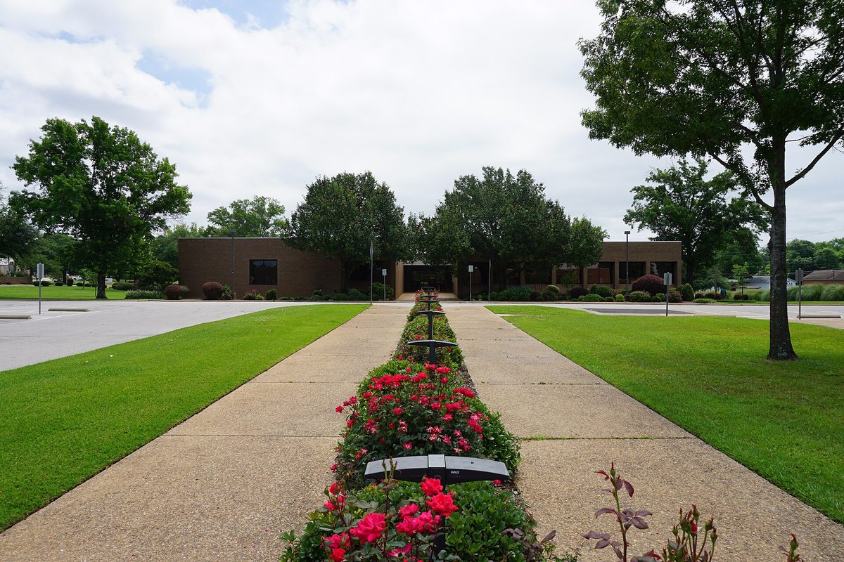 A long concrete walkway lined with red roses leading across green lawns to the low brown brick Longview City Hall, shaded by tall trees