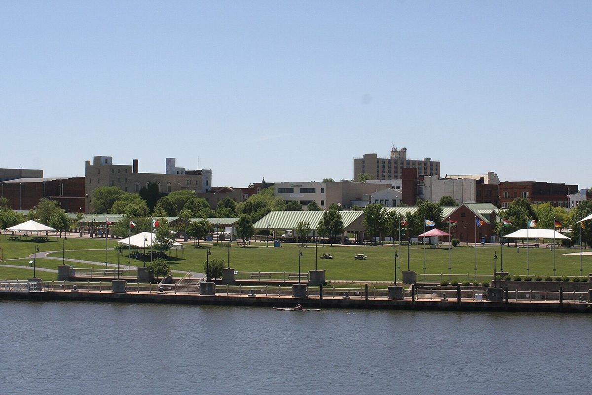 Downtown Lorain seen across the water, with a lawn, white event tents and lamp posts along the riverfront and brick and stone buildings behind