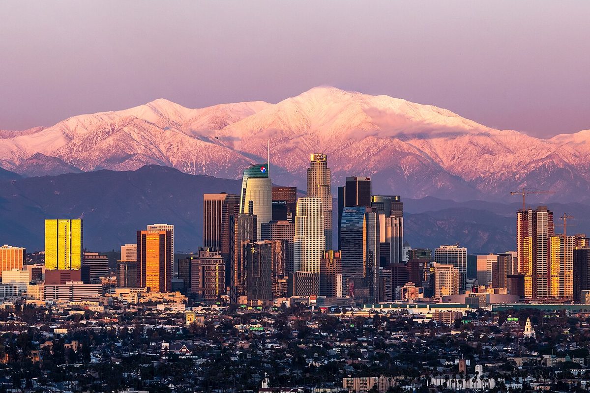 Downtown Los Angeles towers lit by sunset, with snow-covered Mount Baldy rising behind them under a pink sky