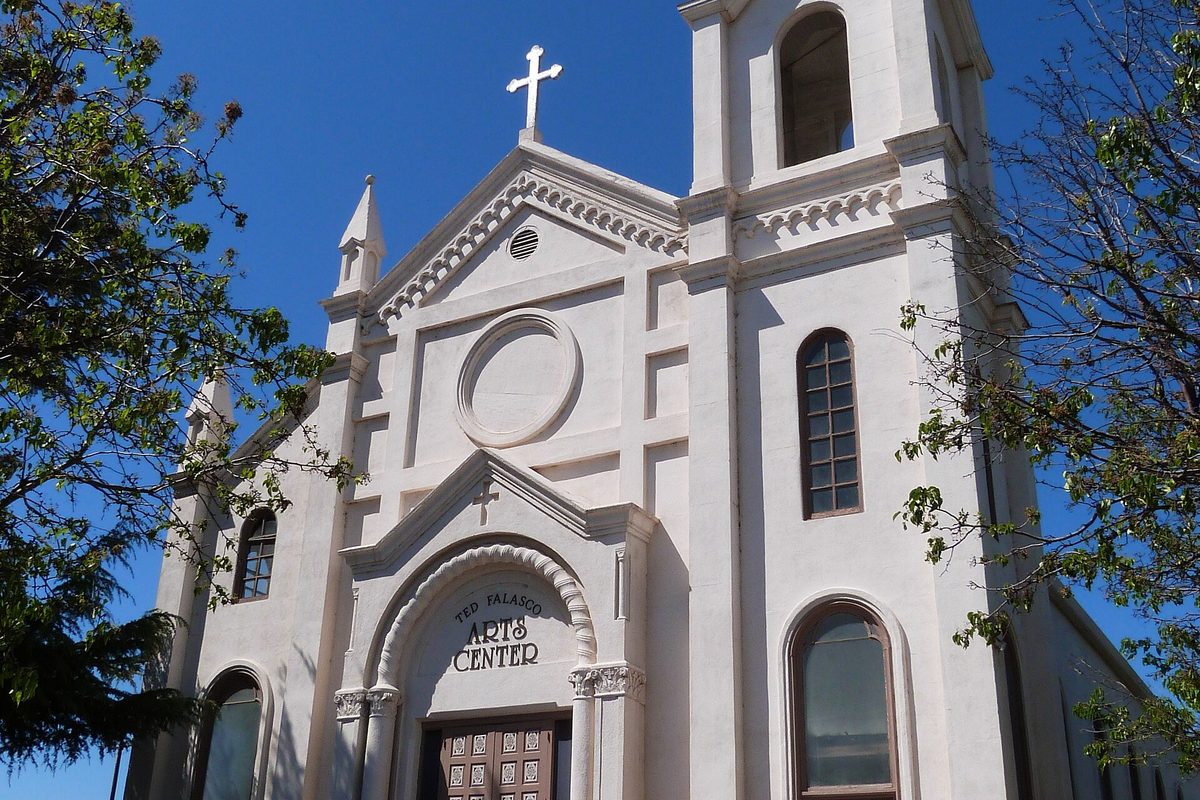 The white facade of the old St. Joseph Church in Los Banos, a cross on top and a sign over the door reading Ted Falasco Arts Center, framed by green trees