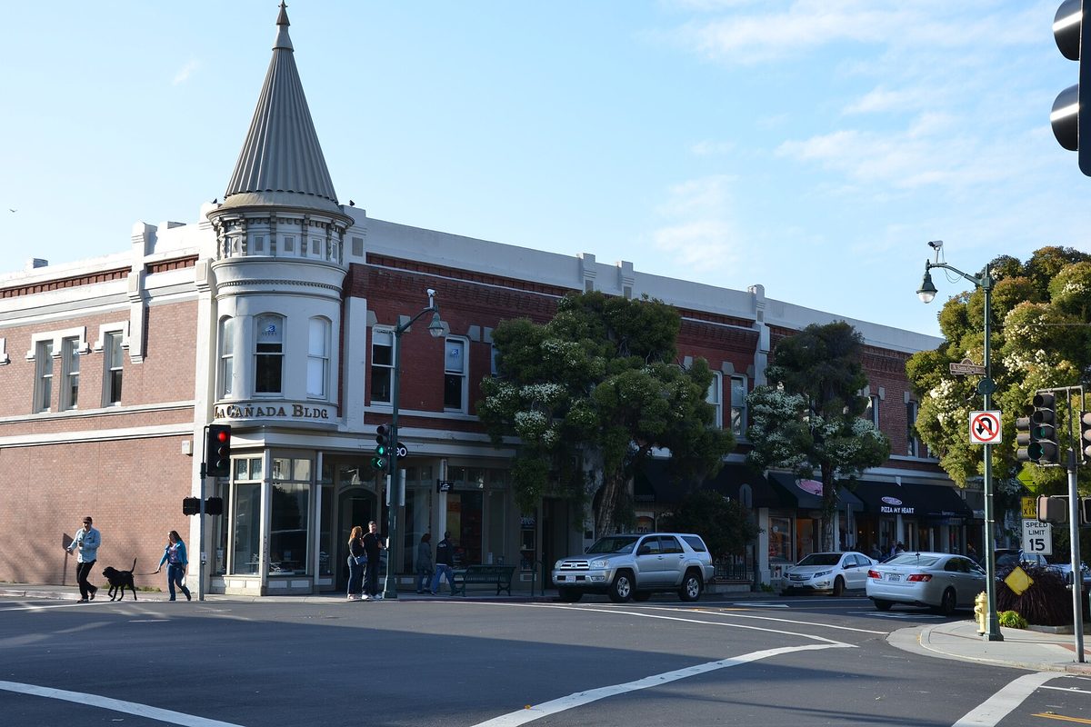 The brick La Cañada Building with its pointed corner turret on a downtown Los Gatos street, with people walking a dog past parked cars