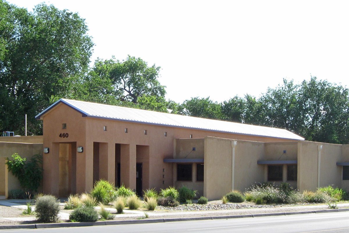 The Los Lunas Public Library at 460 Main Street NE, a low tan stucco building with a metal roof and desert plantings, framed by tall trees