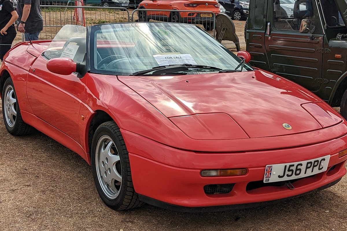 A red Lotus Elan M100 convertible with the top down and silver wheels, a for-sale sign on the windshield, parked on dirt