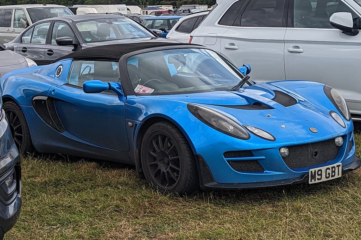 A bright blue Lotus Elise roadster with a black soft top and black wheels, parked on grass among other cars
