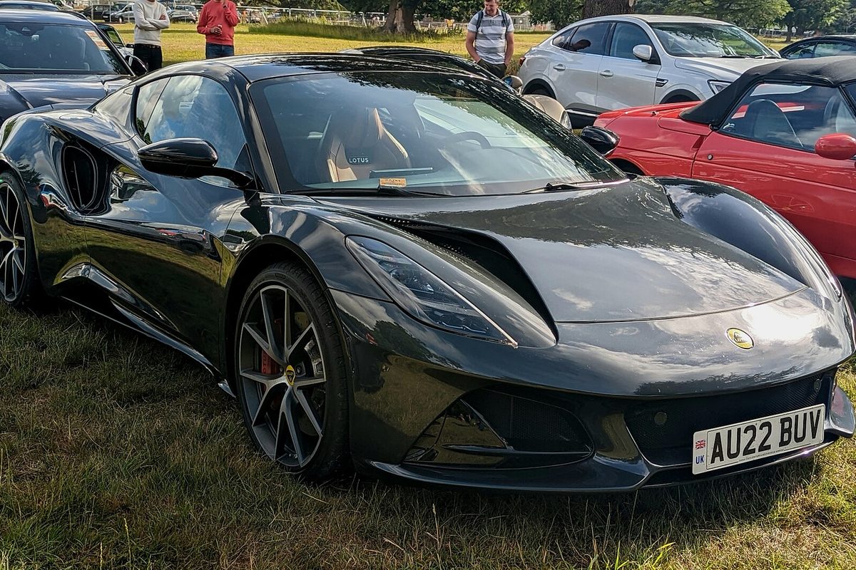 A dark gray Lotus Emira coupe with multi-spoke wheels parked on grass, a red car beside it