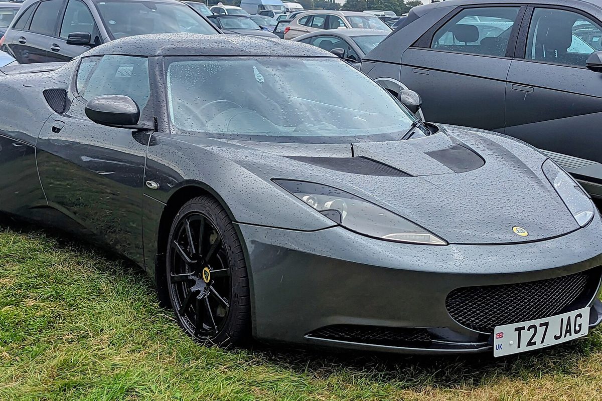 A dark gray Lotus Evora coupe with black wheels, beaded with rain, parked on grass beside other cars