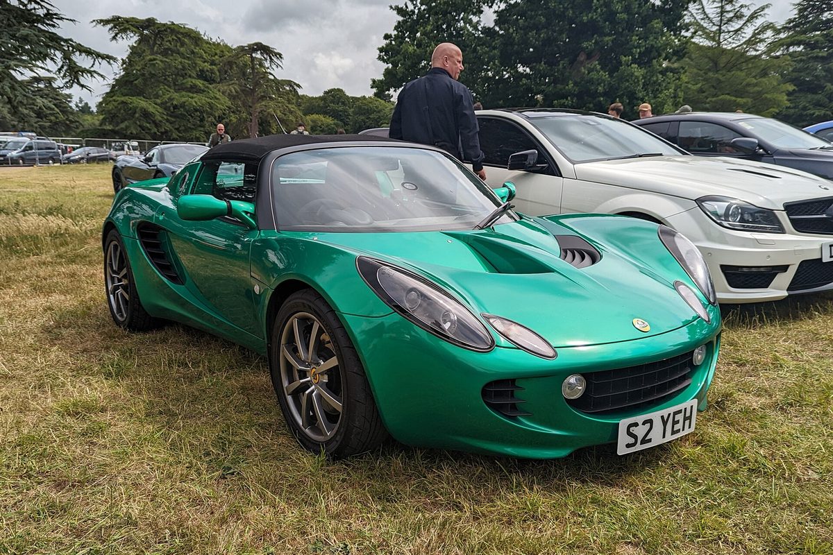 A green Lotus Elise roadster with its black soft top up, parked on the grass at an outdoor car show beside a white sedan