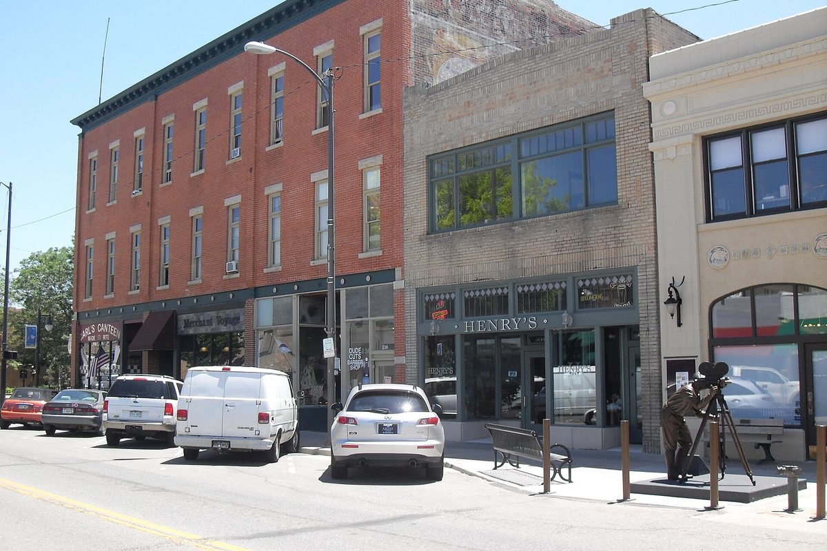 Brick storefronts on 4th Street in downtown Loveland, with cars parked at the curb and a bronze statue of a man at a camera on a tripod on the sidewalk
