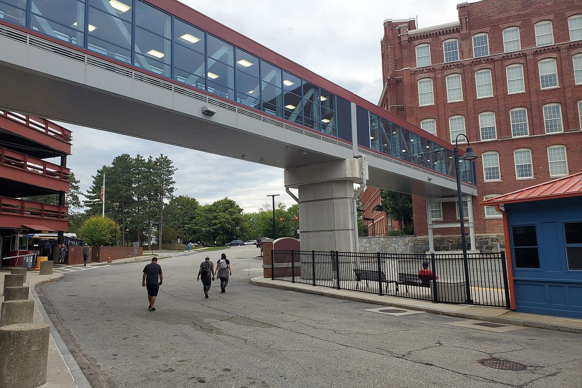 The glass-enclosed footbridge over the road at Lowell's Gallagher Terminal, with a red brick mill building behind it and people walking underneath