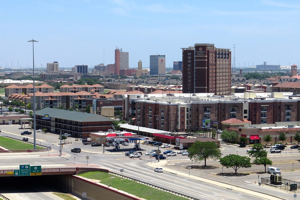 Lubbock's skyline seen from the Marsha Sharp Freeway, with a tall brick hotel tower, low apartment blocks, a parking lot and a green highway sign over an underpass