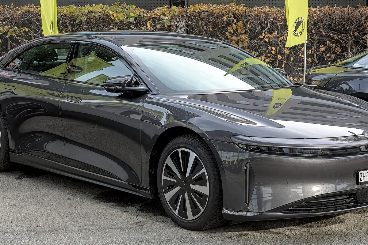 A dark gray Lucid Air sedan with aero wheels, parked on pavement in front of a hedge and yellow flags