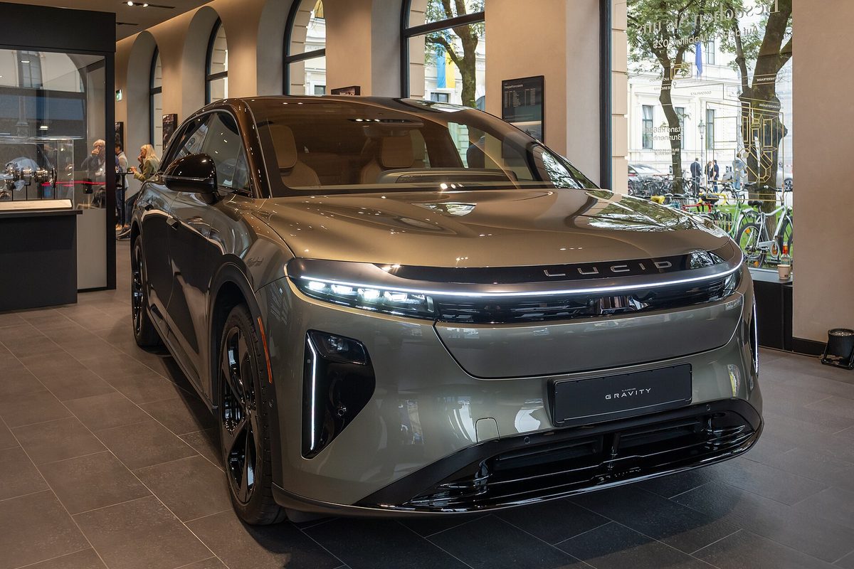A pale green metallic Lucid Gravity SUV with its front light bar lit, parked inside a showroom with tall arched windows