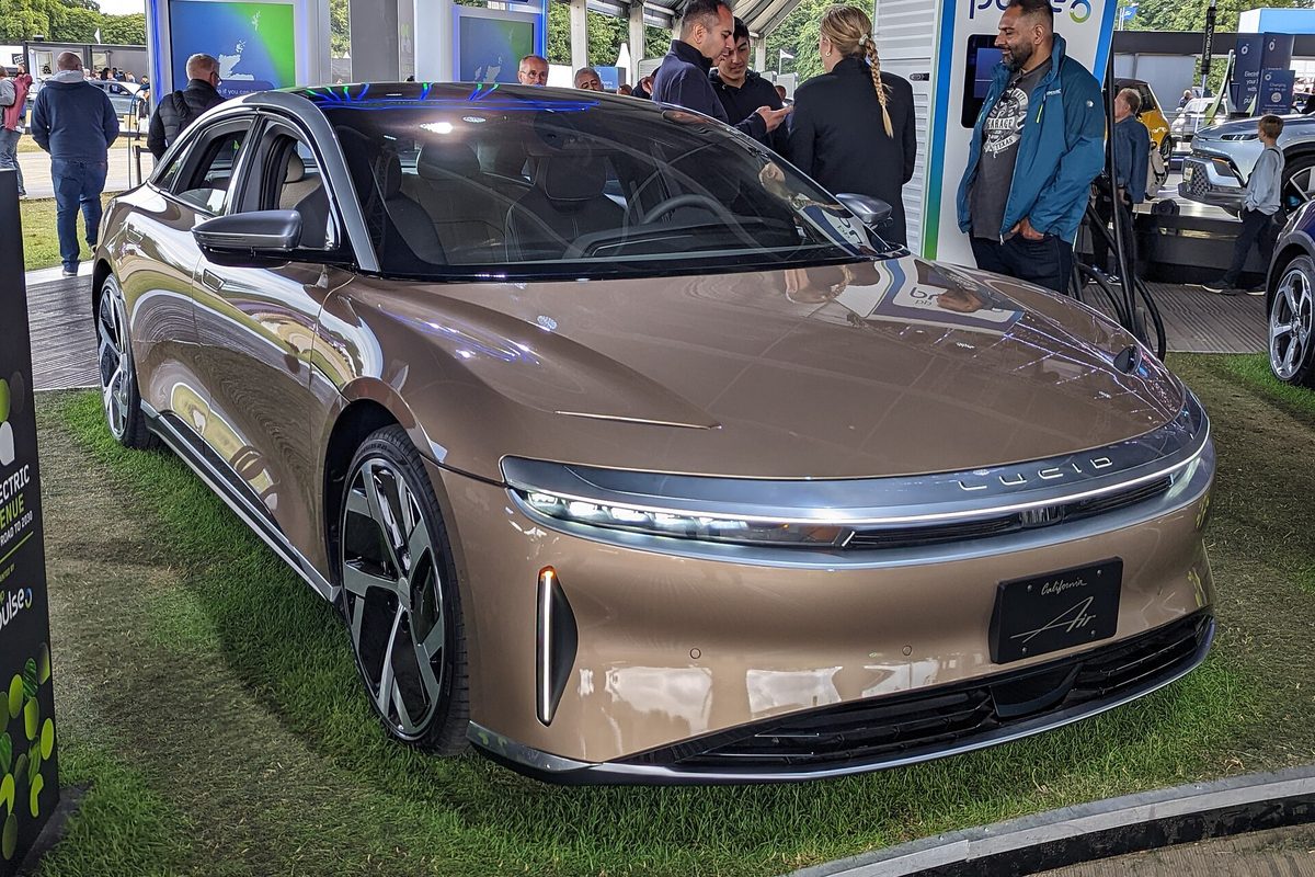 A bronze-colored Lucid Air sedan parked on grass at an outdoor electric vehicle show, with visitors standing behind it