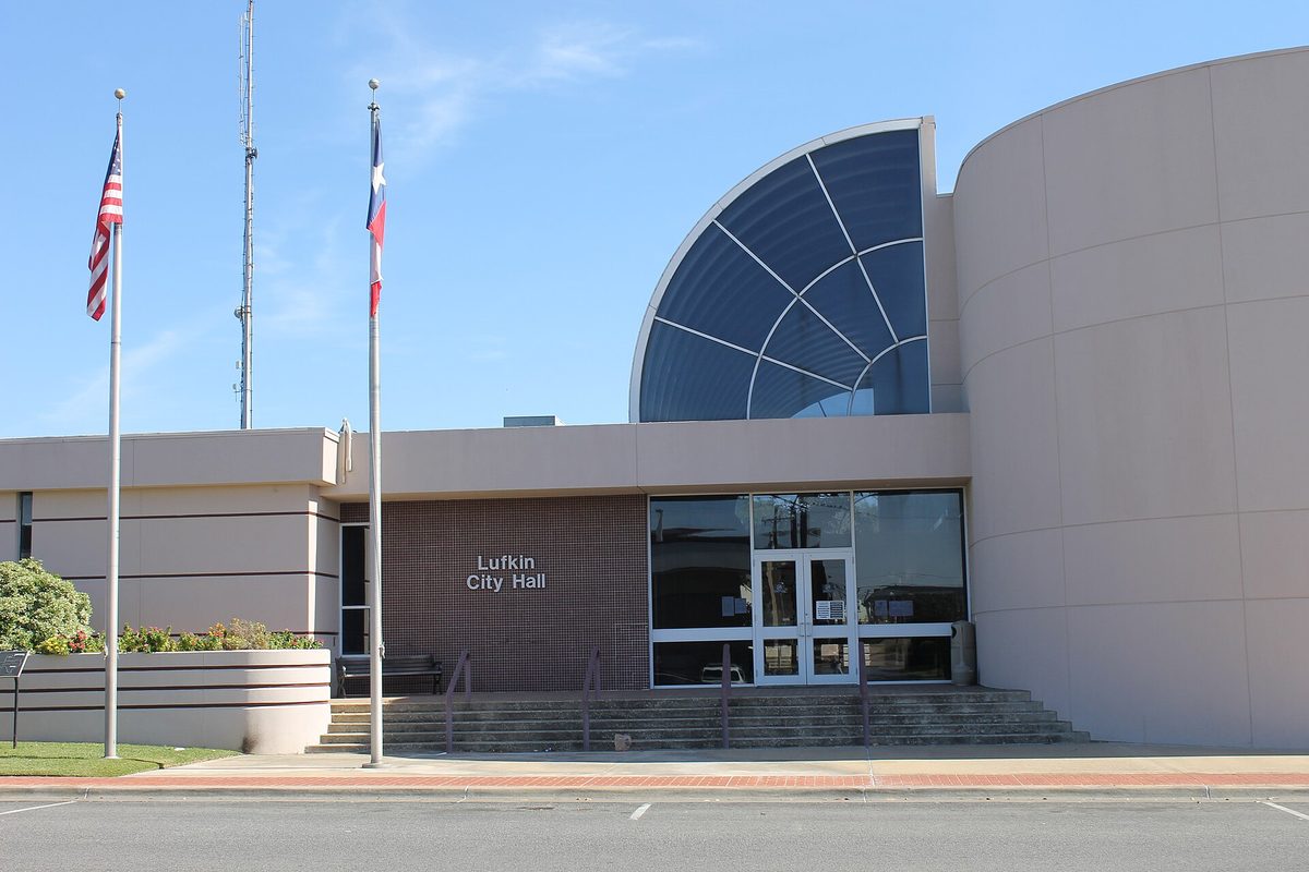 Lufkin City Hall, a tan building with a tall curved glass window over its entrance, with American and Texas flags on poles out front