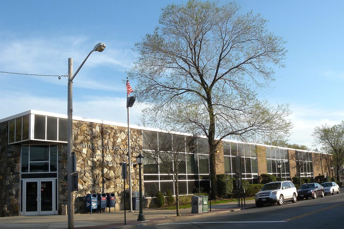 The low stone and glass Lynbrook post office on Broadway at Eldert Street, with a flagpole and a tall bare tree out front on a sunny afternoon