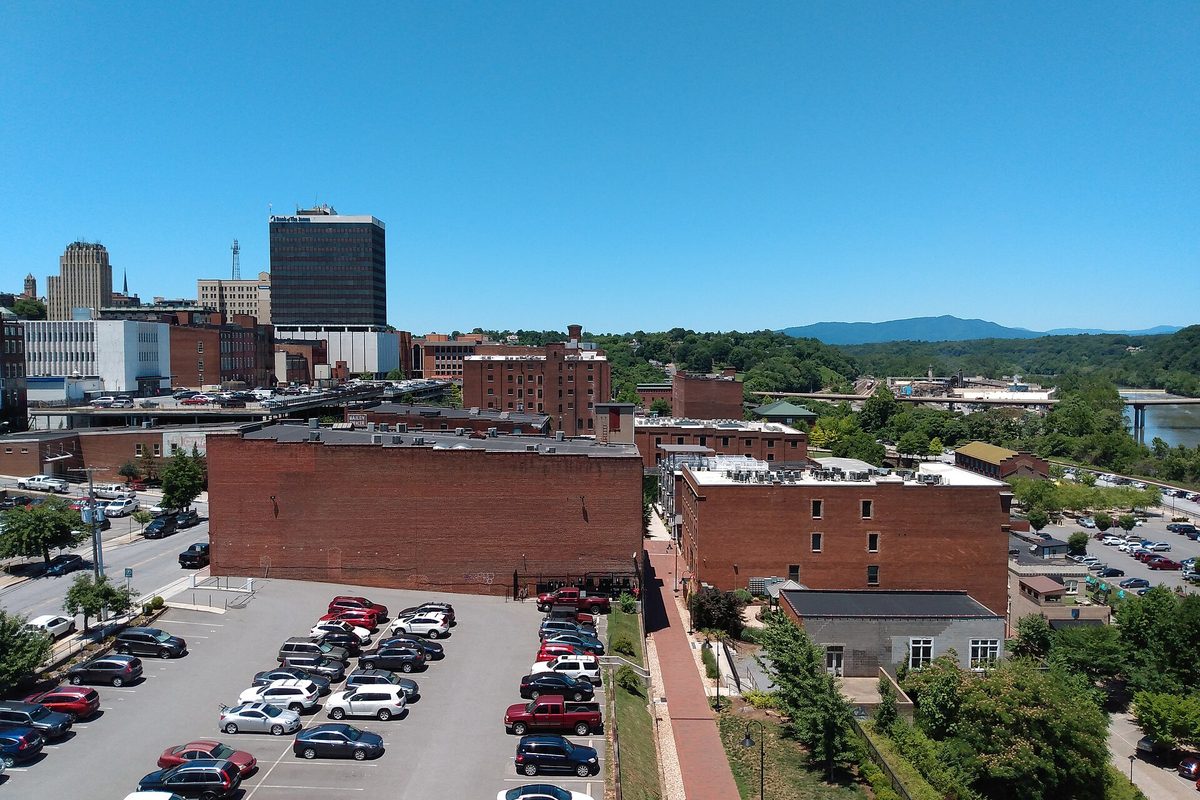 Aerial view of downtown Lynchburg's brick buildings and a full parking lot, with a river, a bridge and a distant mountain under a clear sky