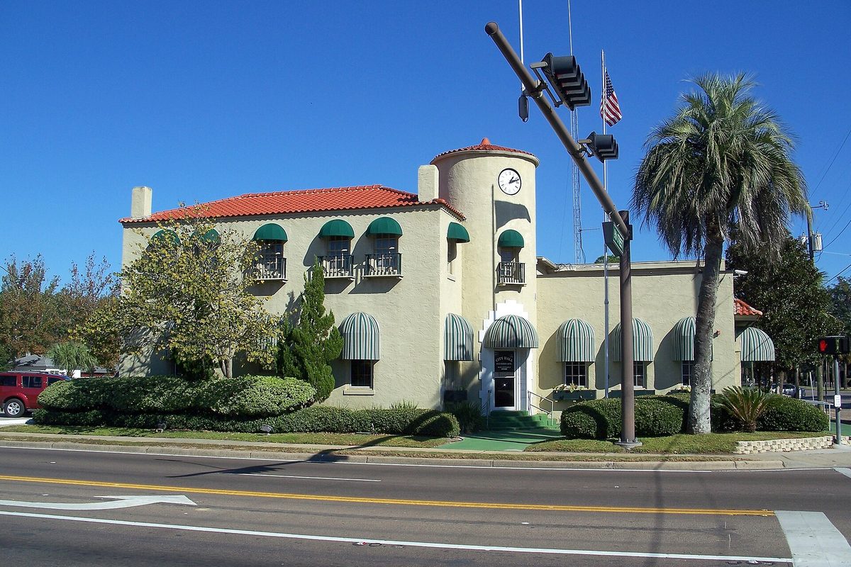 Lynn Haven City Hall, a cream two-story building with a red tile roof, green striped awnings and a round clock tower, beside a palm tree
