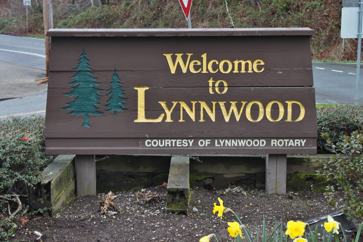 A brown wooden Welcome to Lynnwood sign with painted evergreen trees and the words Courtesy of Lynnwood Rotary, with yellow daffodils in front