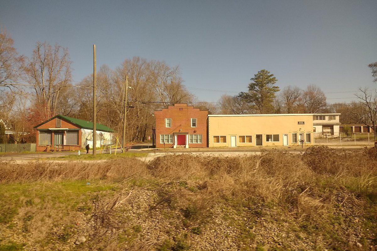 A row of small older buildings in Mableton: a red brick front with a red door between a green-roofed house and a tan block building, behind dry brush and gravel
