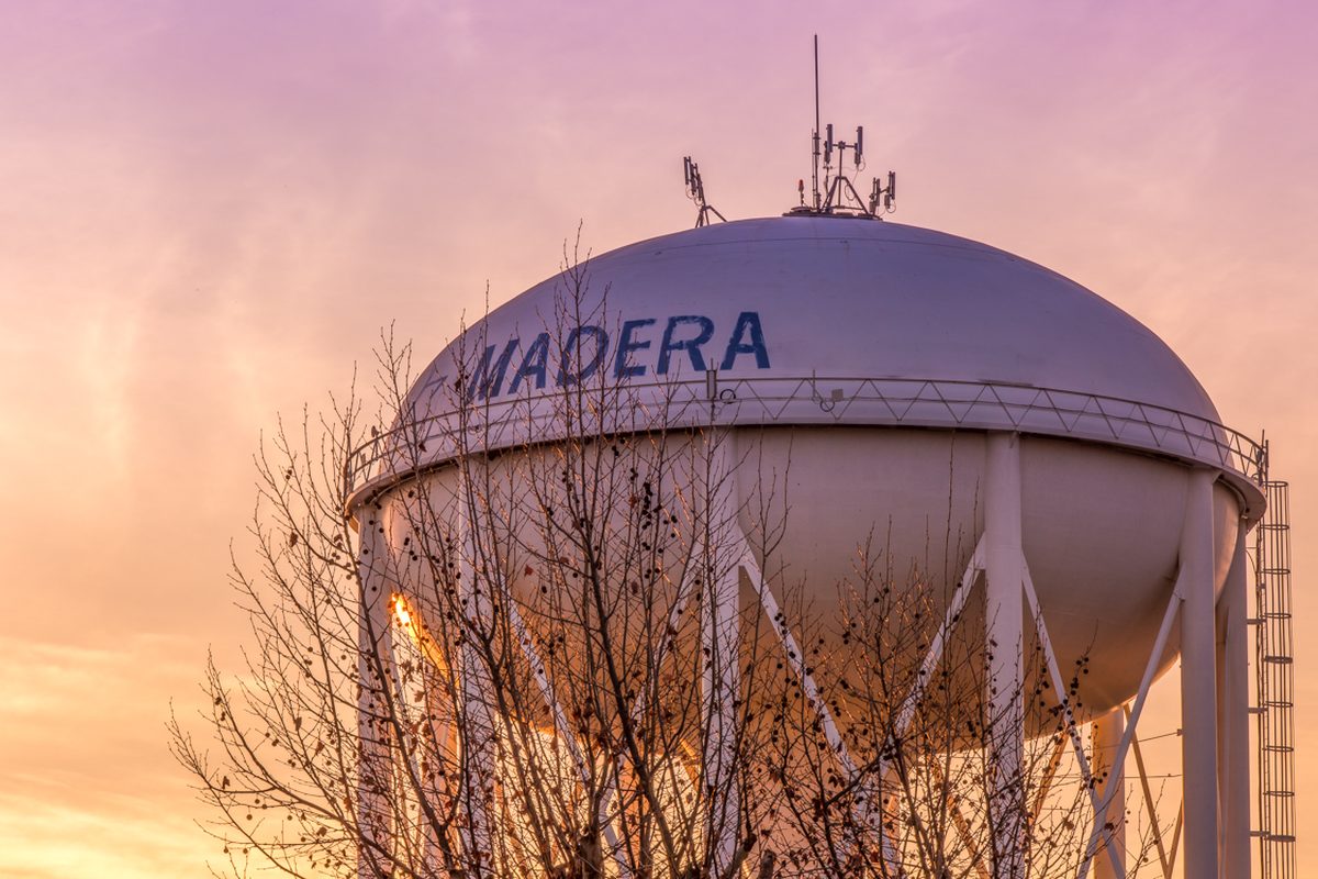 The white City of Madera water tower with the city's name in blue letters, seen past bare tree branches against a pink and orange sunset sky