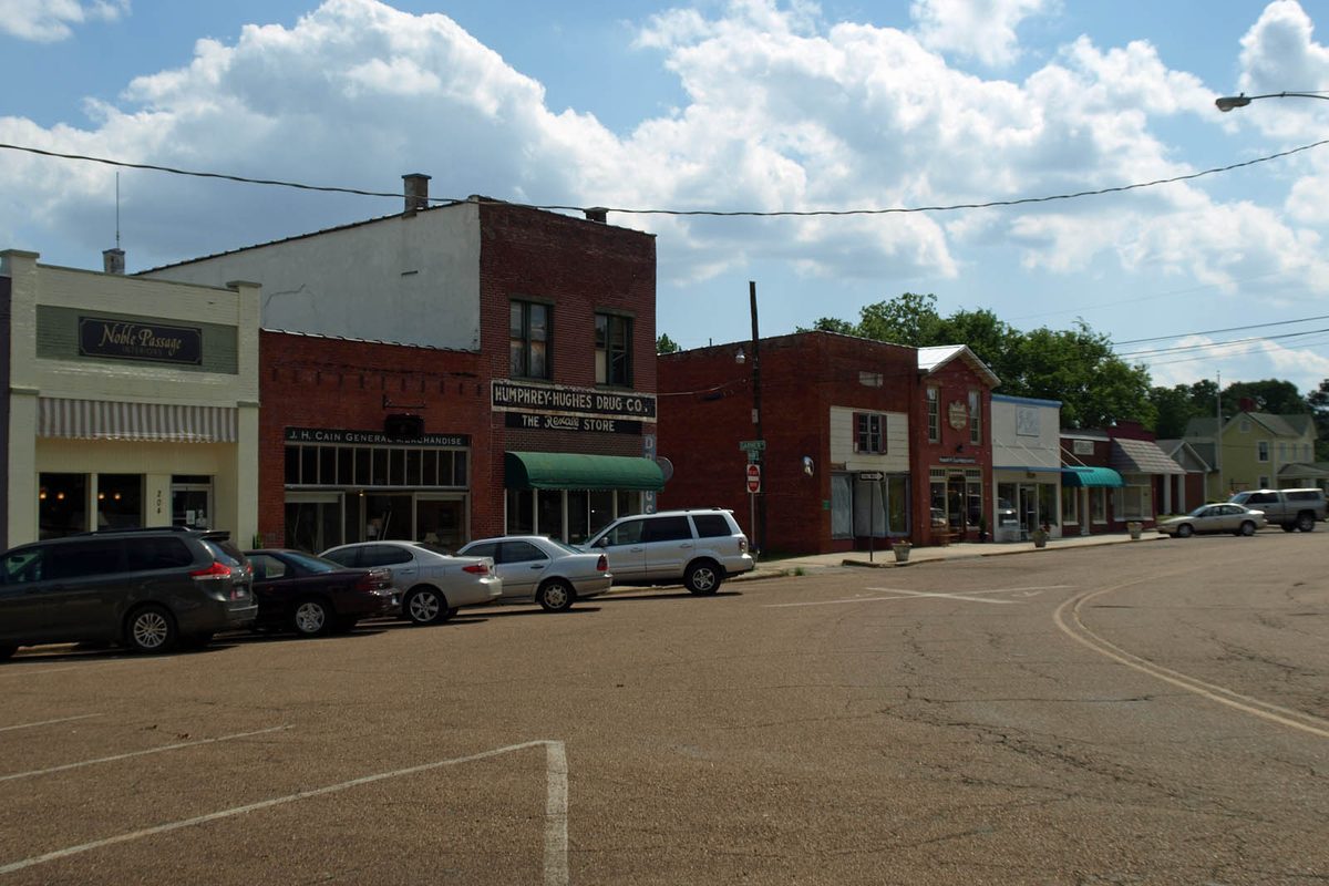 Two-story red brick storefronts with green awnings along Main Street in Madison, with cars parked at the curb under a cloudy sky