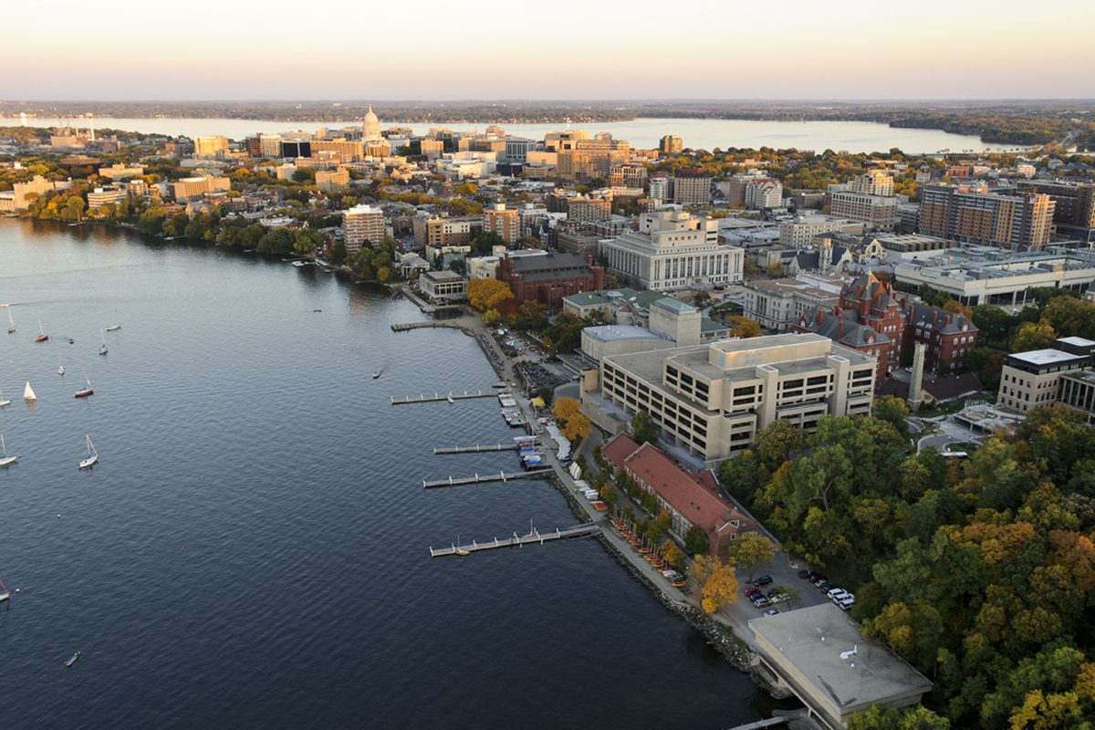 Aerial view at sunset over the UW-Madison campus and the Lake Mendota shoreline, with sailboats and piers in front and the Capitol dome rising beyond downtown